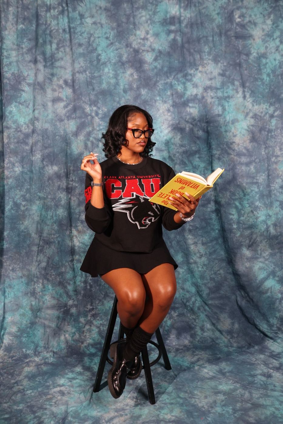 Aja on a stool against a blue backdrop, reading a book held upside down