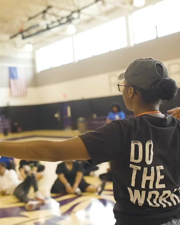A DOTHEWORK crew member leads a group of seated participants in a gym