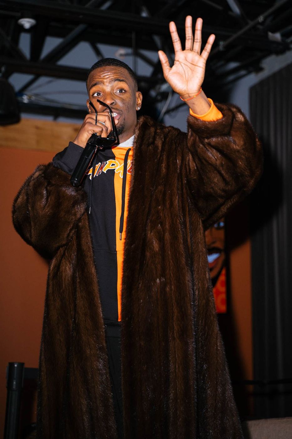 Hail Zel on the mic in a floor-length fur coat, one hand raised to the room