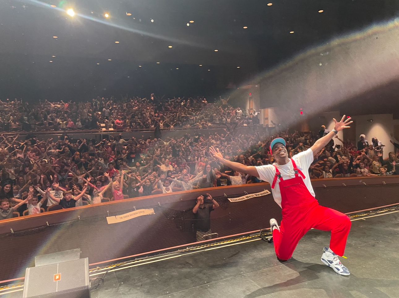 Mr. Prather on one knee onstage, arms wide, in front of a packed house of children with their arms in the air